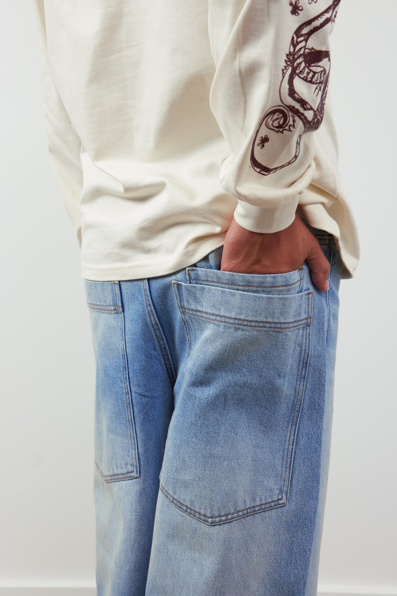 Person wearing a cream long-sleeve shirt with a graphic design and blue jeans on a white background