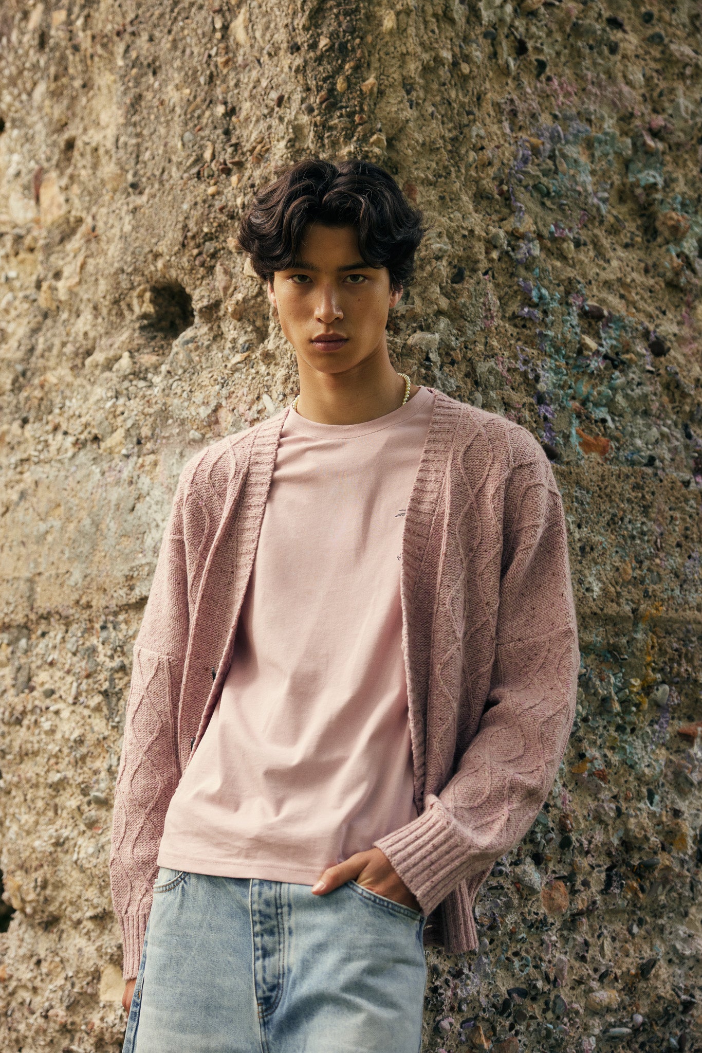 Person wearing a pink cardigan over a light pink shirt with light blue jeans, standing against a textured wall.
