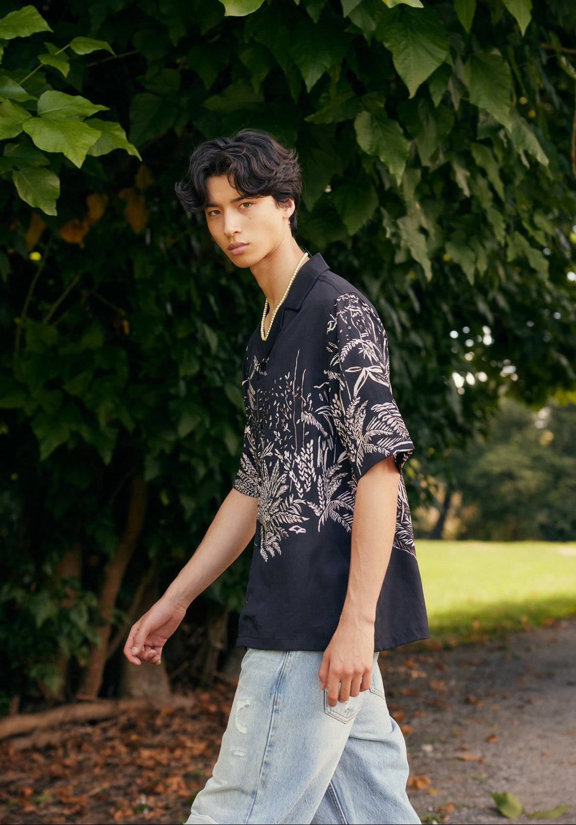 Person wearing a dark floral shirt and light jeans standing in a natural setting with greenery.