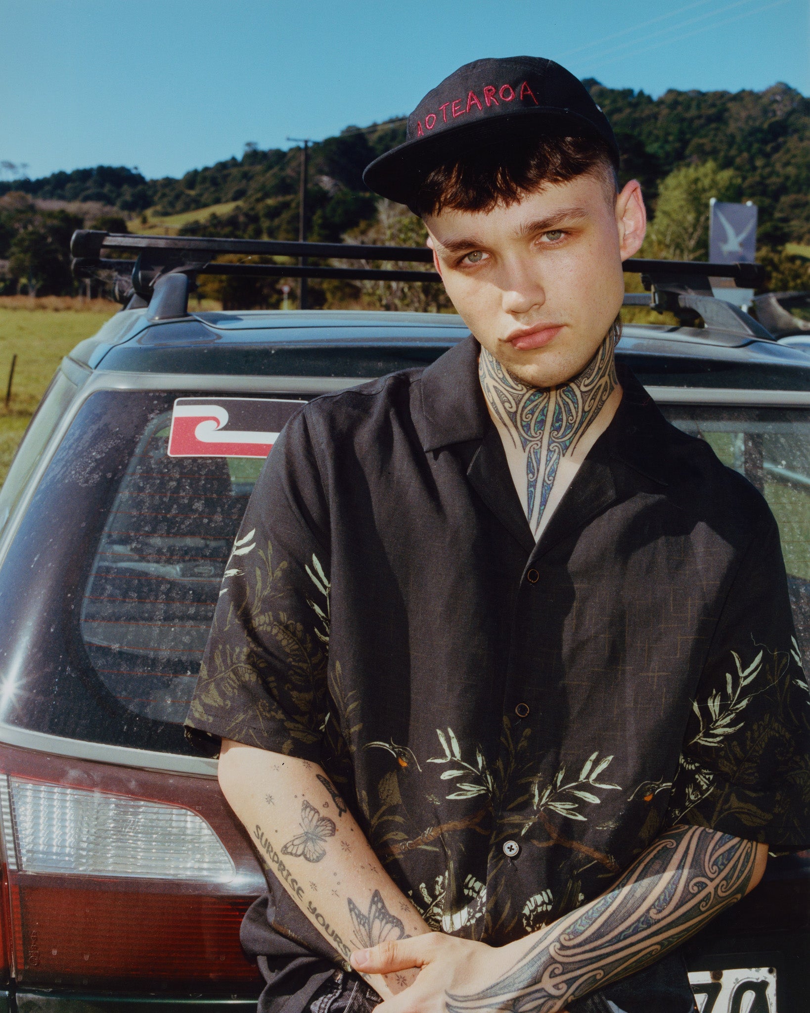Person with tattoos standing in front of a car with a scenic background