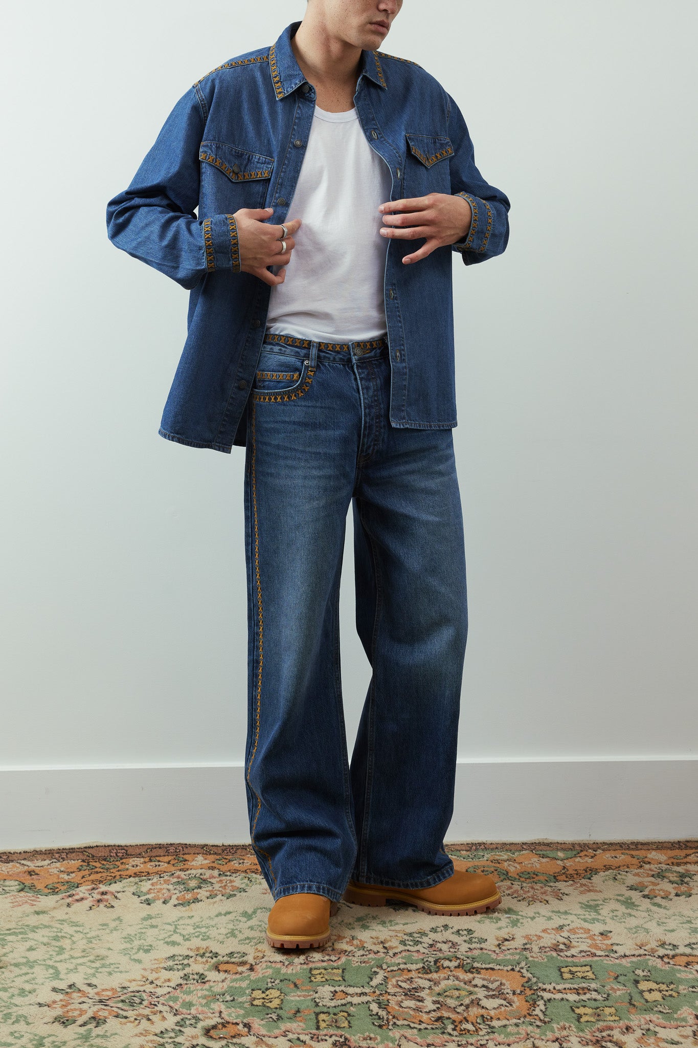 A model wearing a blue denim shirt jacket over a white t-shirt, paired with wide-leg blue jeans and tan boots, standing on a patterned rug against a light gray wall.