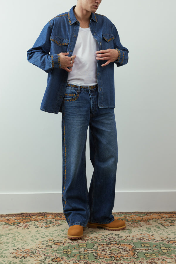 A model wearing a blue denim shirt jacket over a white t-shirt, paired with wide-leg blue jeans and tan boots, standing on a patterned rug against a light gray wall.