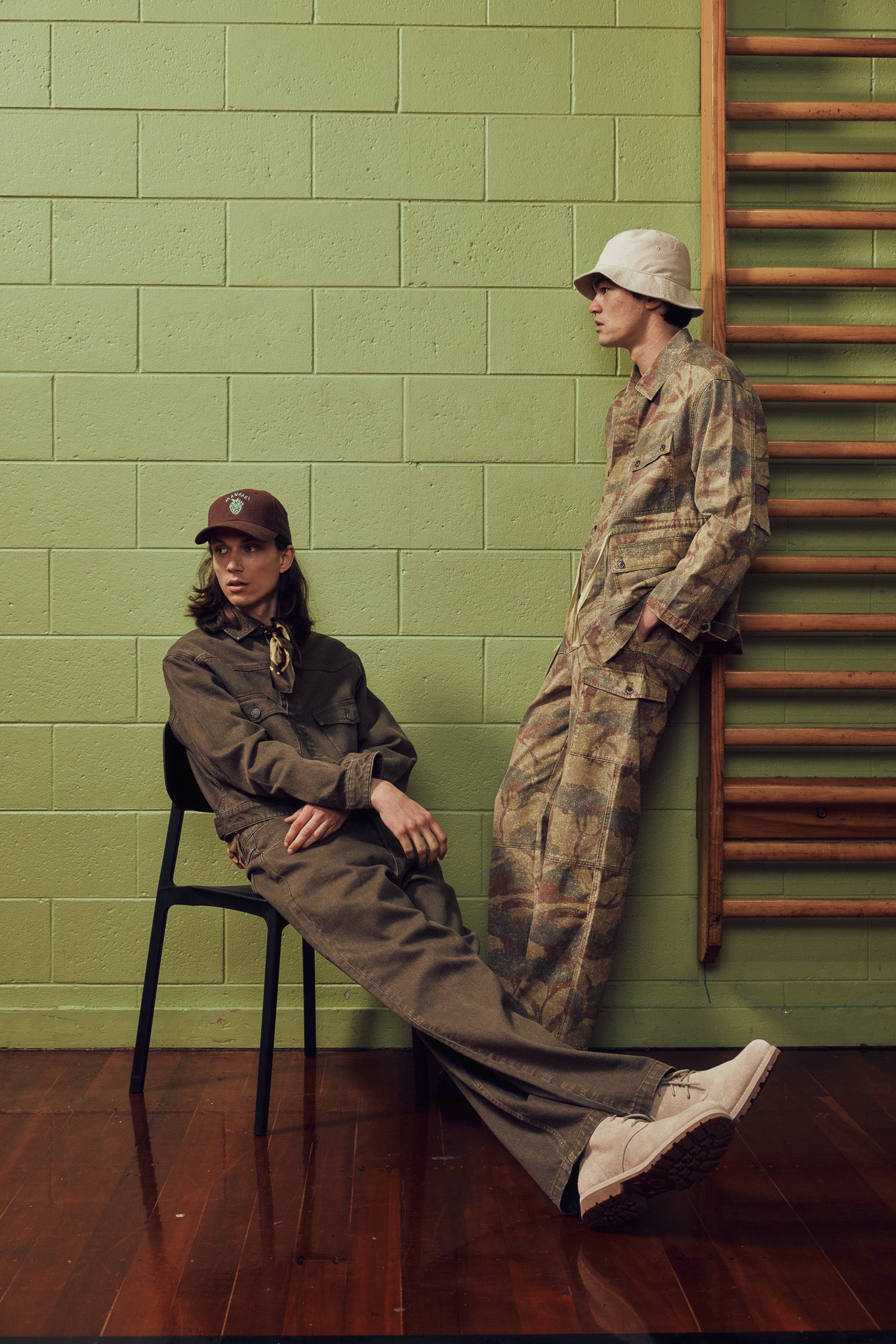 Two models posing in military-style jumpsuits against a green brick wall. One model sits on a black chair wearing a dark olive-brown jumpsuit and a brown cap, while the other stands leaning against a wooden ladder wearing a camouflage jumpsuit and a white bucket hat.
