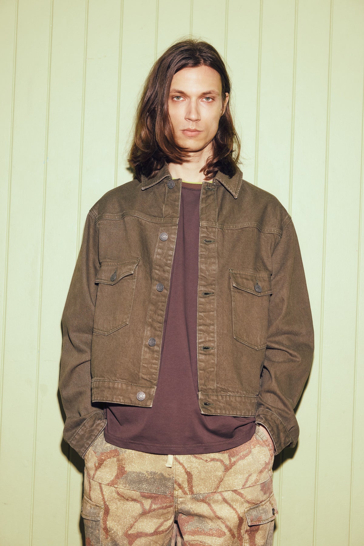 A model with long brown hair stands against a pale green paneled wall, wearing a brown corduroy utility jacket over a dark brown t-shirt and beige patterned cargo pants.