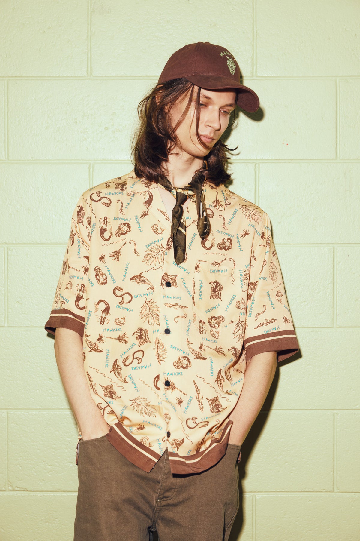 A person wearing a brown baseball cap, a beige short-sleeve button-up shirt with a brown and blue graphic print, and brown pants, standing against a pale green painted brick wall.