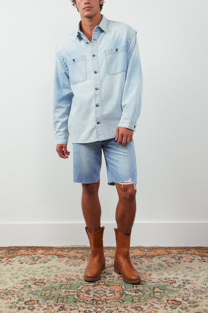 Person wearing a light blue denim shirt, shorts, and brown boots on a white background