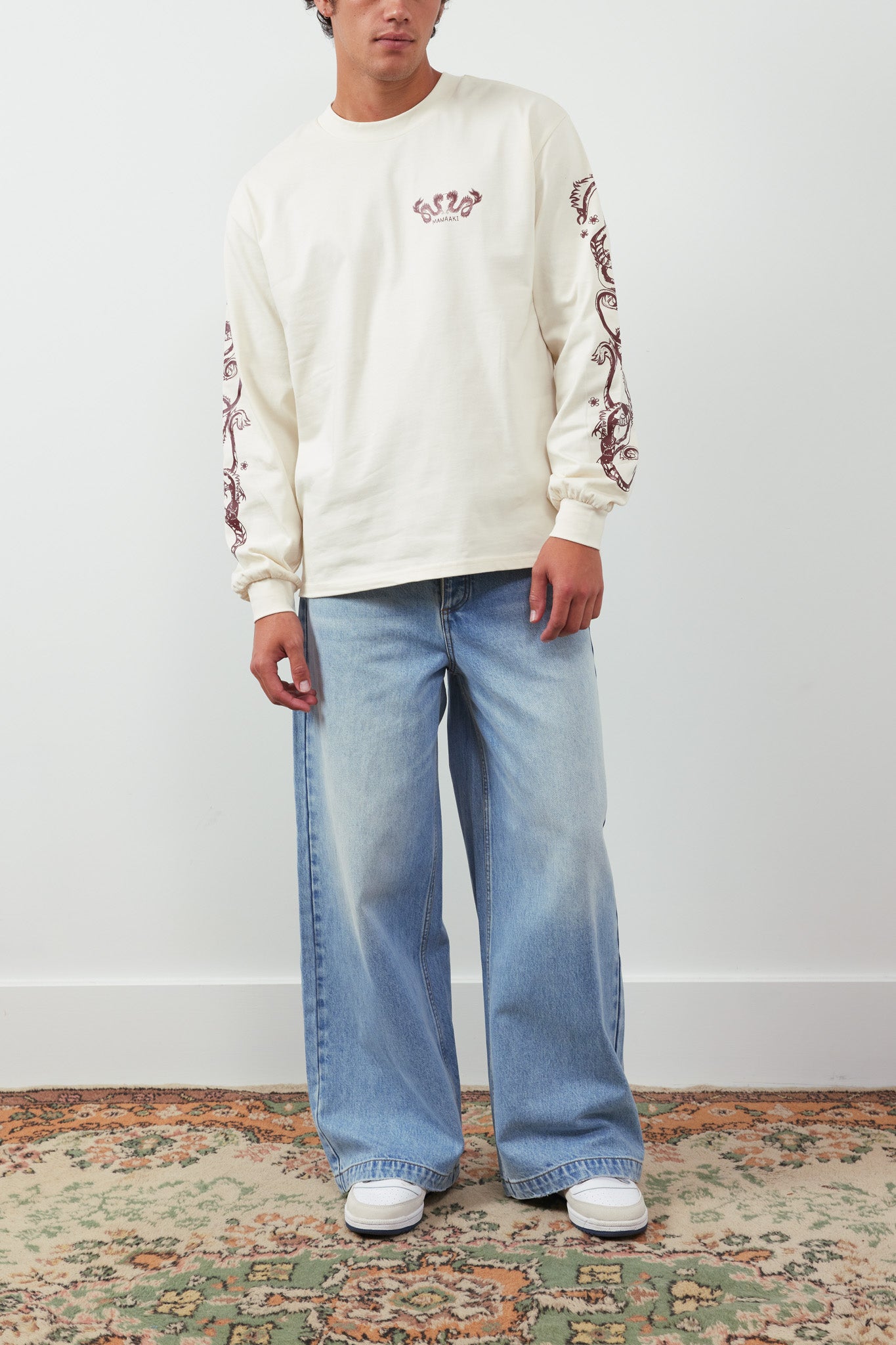 Person wearing a cream sweatshirt with floral patterns and light blue jeans on a plain background