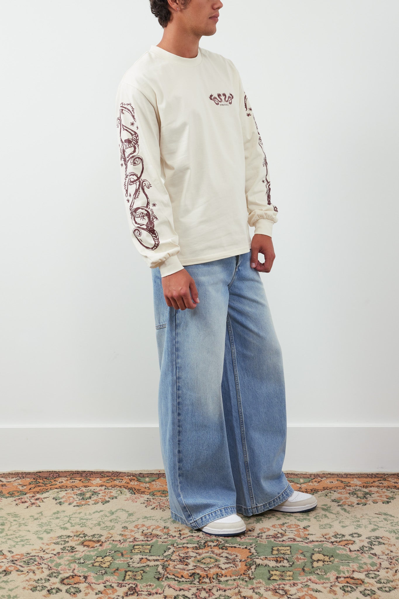 Person wearing a cream sweatshirt with red designs and light blue jeans on a white background