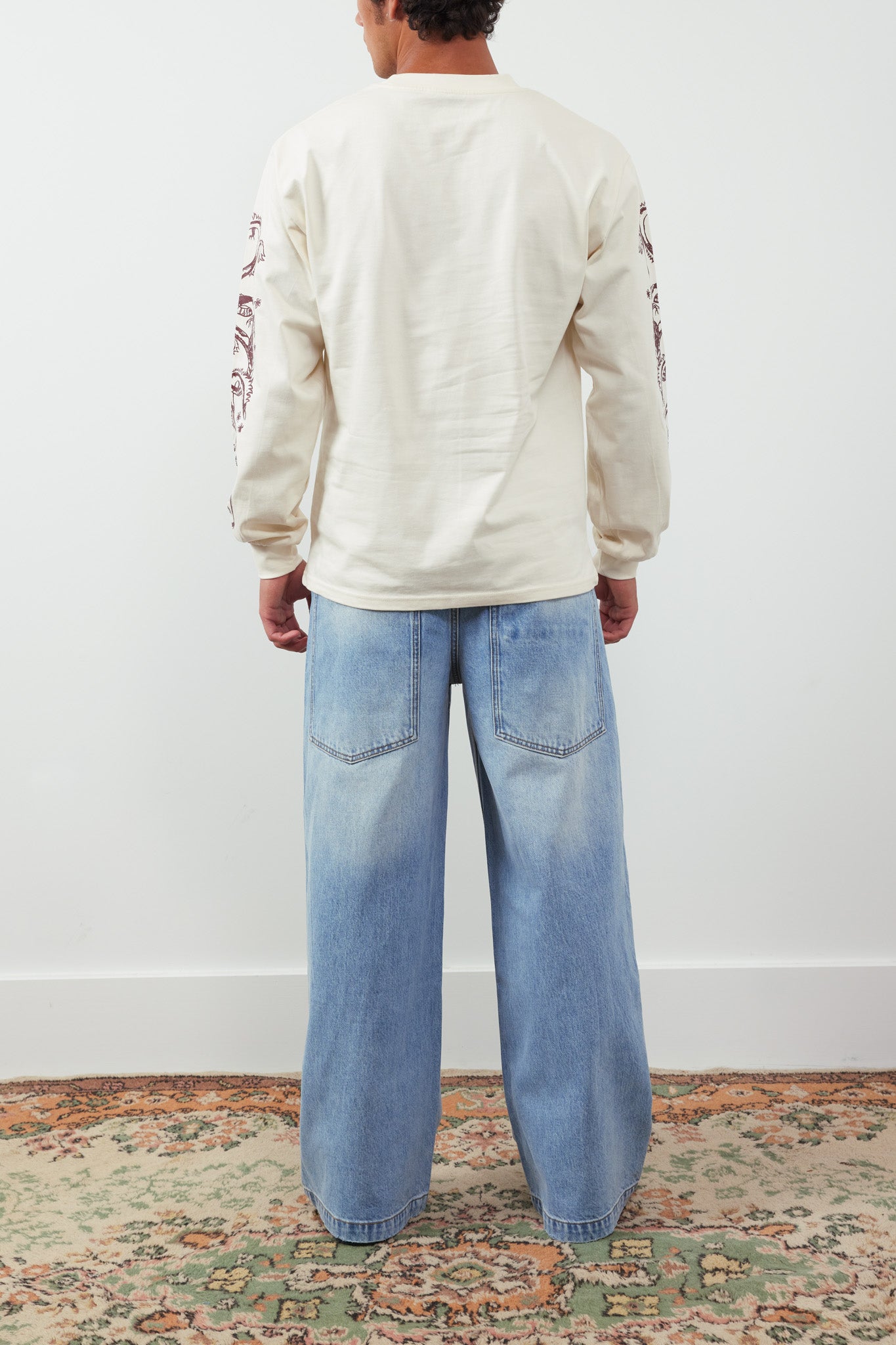 Person wearing a cream sweatshirt and light blue jeans standing on a patterned rug.