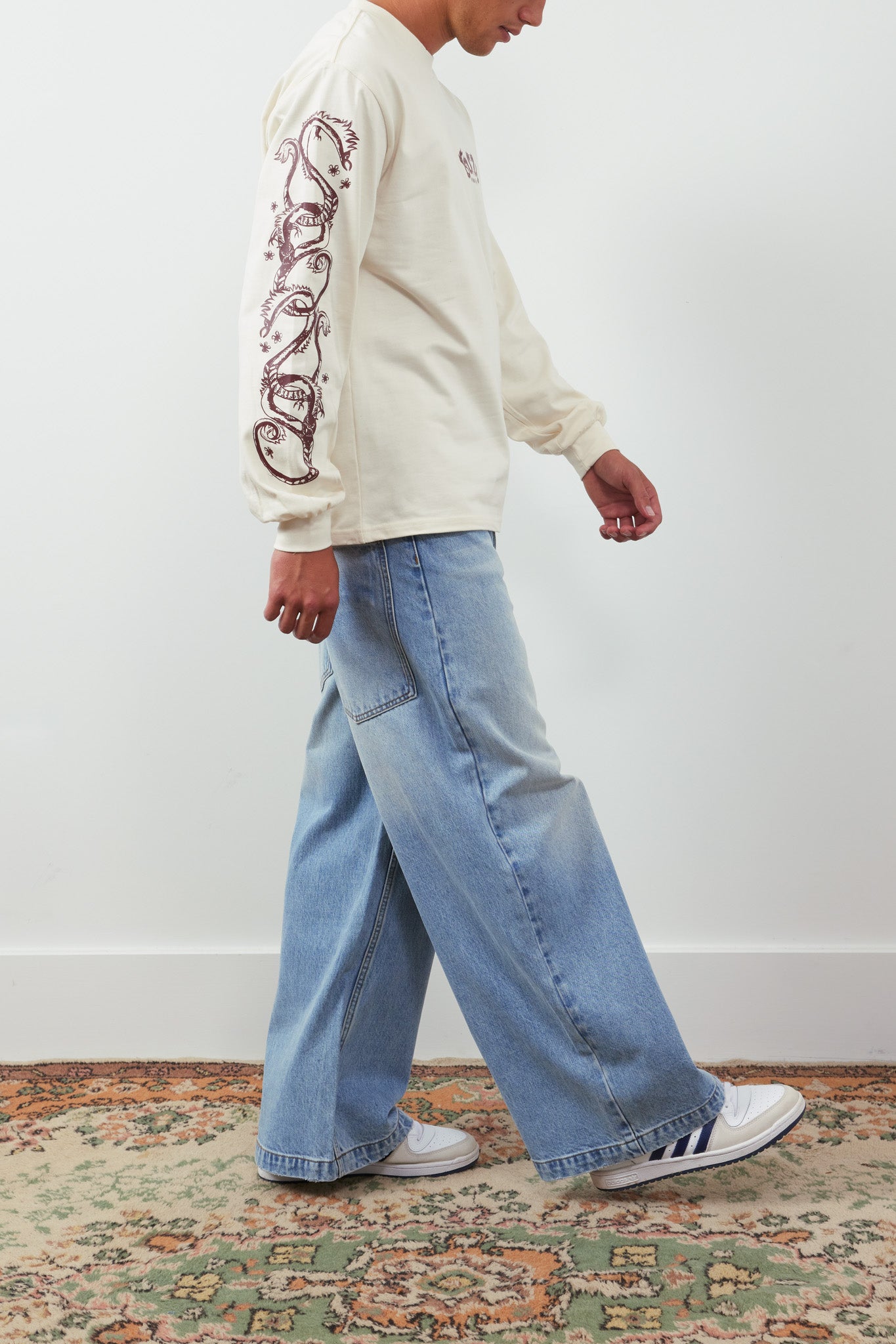 Person wearing a cream sweatshirt with intricate designs and blue jeans, walking on a patterned rug.