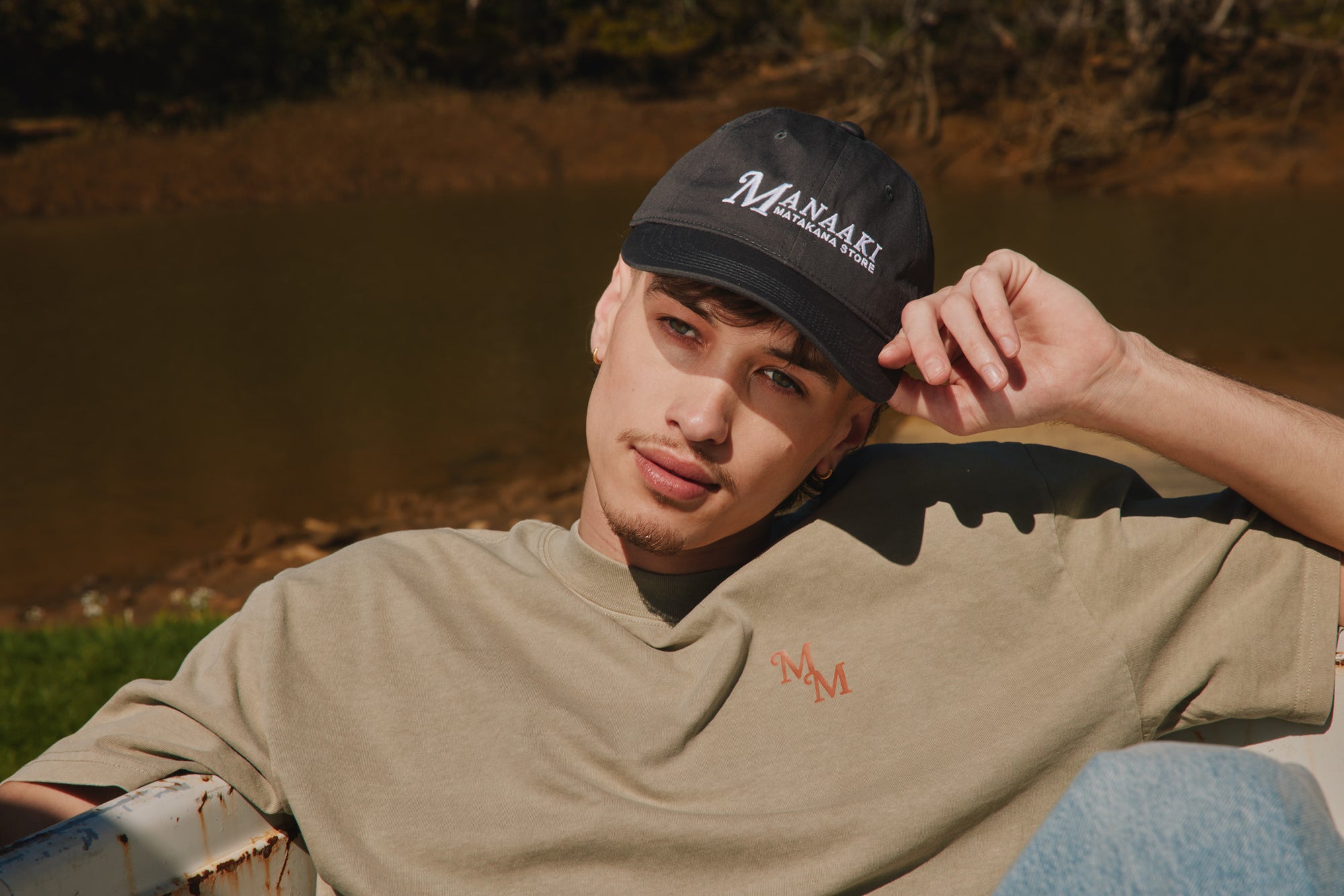 Person wearing a cap and t-shirt with a logo, sitting outdoors by a body of water.