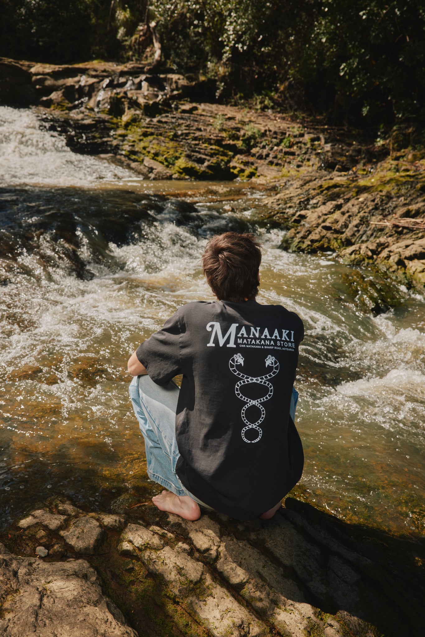Person wearing a black shirt with 'Manaaki' branding sitting by a flowing stream.