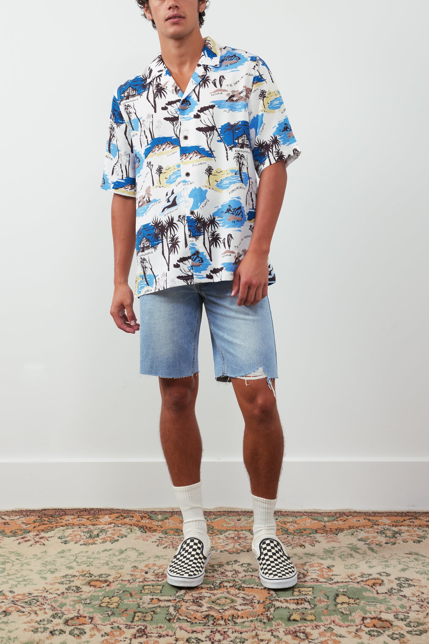 Person wearing a colorful floral shirt and denim shorts standing on a patterned rug.